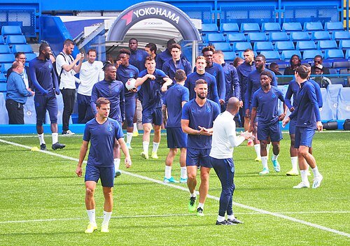 Chelsea's squad training on the stadium pitch, players in blue kit with the empty stands in the background