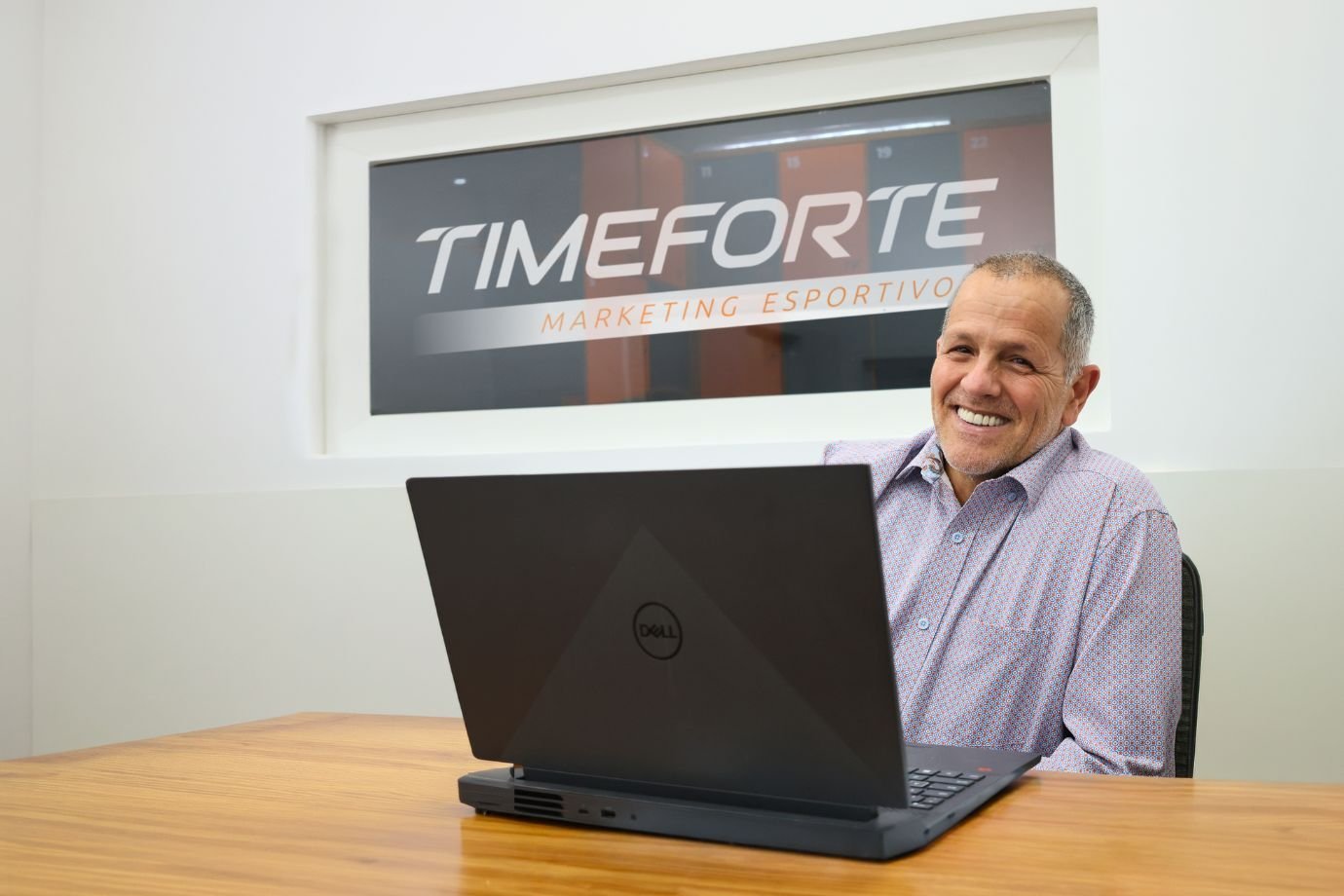 José Martins, founder of Time Forte, smiling at his desk with a laptop, in front of the Time Forte Marketing Esportivo sign on the office wall