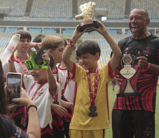 Crianças com medalhas comemoram no gramado do Maracanã, com jovem goleiro erguendo troféu de luva dourada ao lado de um ídolo do Flamengo