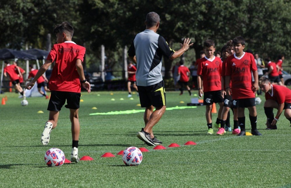 Crianças de camisa rubro-negra treinam com bolas e cones sob orientação de um instrutor no gramado do Flamengo Soccer Camp em Orlando, nos Estados Unidos