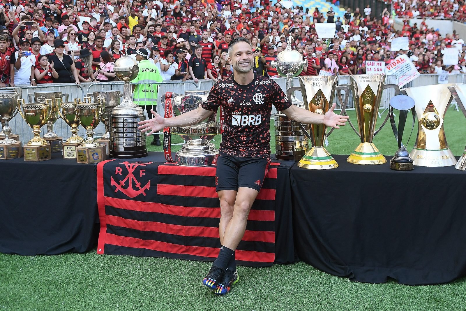Diego Ribas de braços abertos com uniforme do Flamengo diante de mesas com taças do clube no gramado do Maracanã, com torcida ao fundo