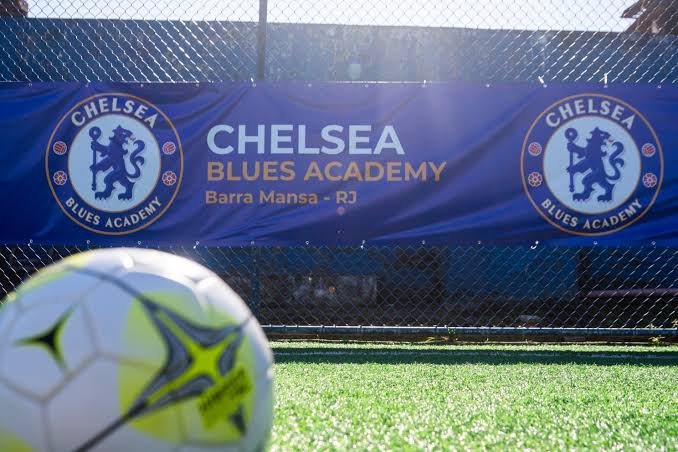 Blue Chelsea Blues Academy banner reading "Barra Mansa - RJ" fixed to the fence of a pitch, with a football in the foreground