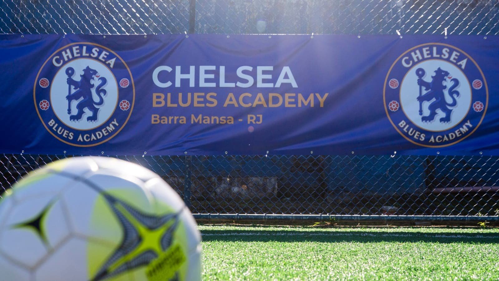 Banner azul da Chelsea Blues Academy com a inscrição "Barra Mansa - RJ" preso ao alambrado de um campo, com uma bola de futebol em primeiro plano sobre a grama
