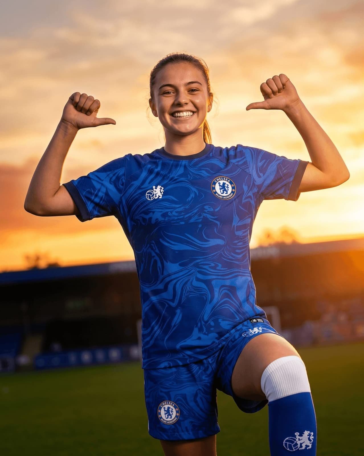 Adolescente da Chelsea Blues Academy uniformizada sorrindo e apontando para o escudo do clube na camisa, com o pôr do sol ao fundo do campo